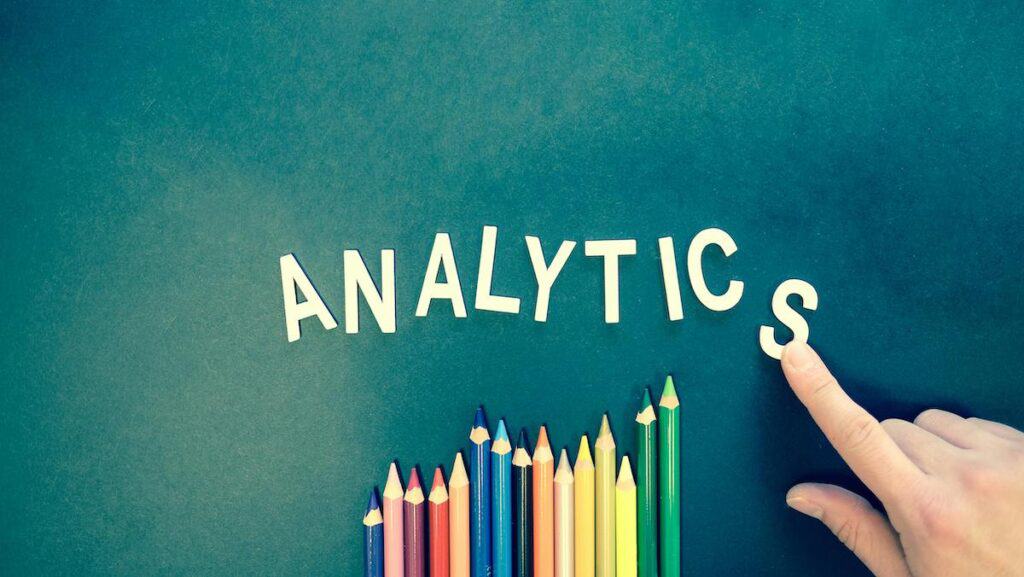 Creative layout with the word 'ANALYTICS' spelled out in white letters against a teal background, with a colorful row of sharpened pencils arranged below. A hand is positioning the letter 'S' at the end of the word.